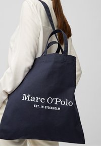 Tmavě modrá plátěná taška přes rameno s dvojitými popruhy. Na jedné straně se nachází bílý text: "Marc O’Polo EST. V STOCKHOLMU". Jednoduchý a funkční design.