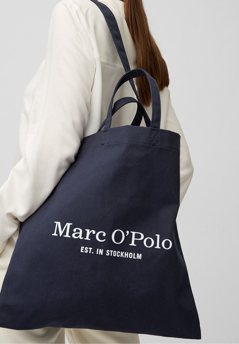Tmavě modrá plátěná taška přes rameno s dvojitými popruhy. Na jedné straně se nachází bílý text: "Marc O’Polo EST. V STOCKHOLMU". Jednoduchý a funkční design.