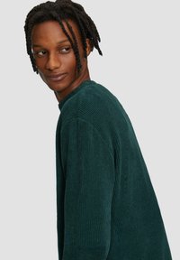 Dunkelgrüner gerippter Pullover mit Rundhalsausschnitt, subtiler Textur und überschnittenen Schultern, in einem lässigen Schnitt gestylt.