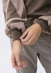 Mains ajustant le poignet d'un pull marron à motifs, portant un pantalon à carreaux avec une grosse boucle de ceinture ronde.