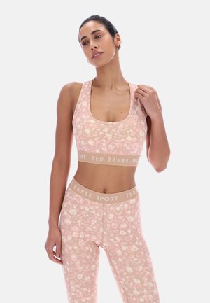 Kvinne iført rosa og beige sports-BH og leggings med blomsterprint, med «Ted Baker Sport» skrevet på midjebåndene, poserer foran hvit bakgrunn.