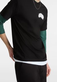 Czarny t-shirt z krótkim rękawem z białym wzorem graficznym na klatce piersiowej, nałożony na tealową koszulę z długim rękawem. Posiada luźny krój.