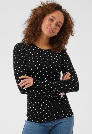 Personne aux cheveux bouclés portant une chemise noire à manches longues à pois blancs et un jean bleu, debout les bras croisés devant un fond uni.