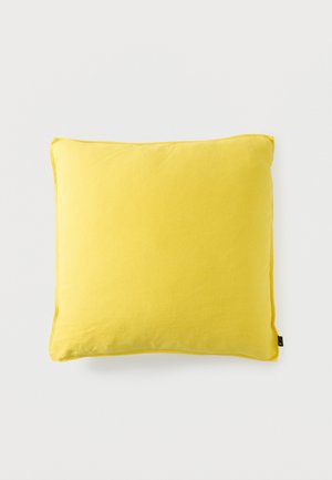 Felgele vierkante kussen met zachte stoftextuur en een klein zwart label aan één rand tegen een effen witte achtergrond.