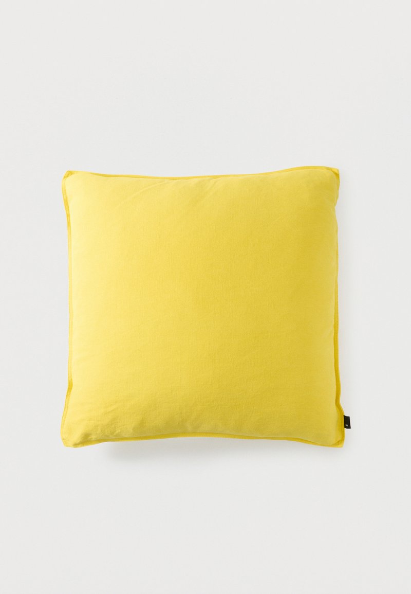 Coussin carré de couleur jaune vif avec une texture de tissu douce et une petite étiquette noire sur un bord, sur un fond blanc uni.