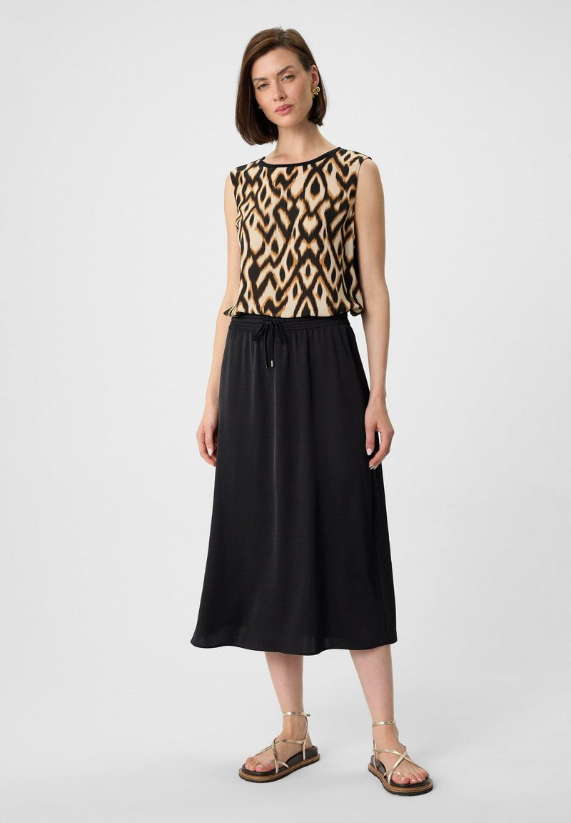 Mouwloze jurk met zwart en beige geometrisch patroon, cinch taille, vloeiende zwarte rok, gecombineerd met sandaaltjes met bandjes.
