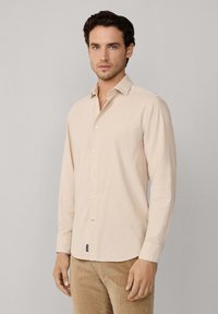 Chemise beige à manches longues avec un col classique, des boutons sur le devant et une texture subtile. Portée avec un pantalon en velours côtelé.