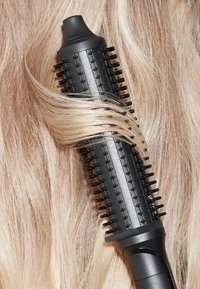 Zwarte verwarmde stylingborstel met een ronde cilinder, rubberen grip en meerdere kleine borstels, die voorzichtig delen van lang, lichtblond haar krullen.