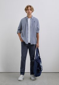 Joven vestido con una camisa a cuadros sobre una camiseta blanca, jeans oscuros, zapatillas blancas, sosteniendo una bolsa de tela azul marino con un suéter doblado dentro.