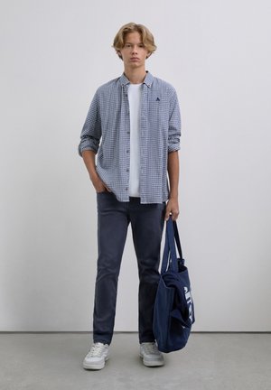 Joven vestido con una camisa a cuadros sobre una camiseta blanca, jeans oscuros, zapatillas blancas, sosteniendo una bolsa de tela azul marino con un suéter doblado dentro.