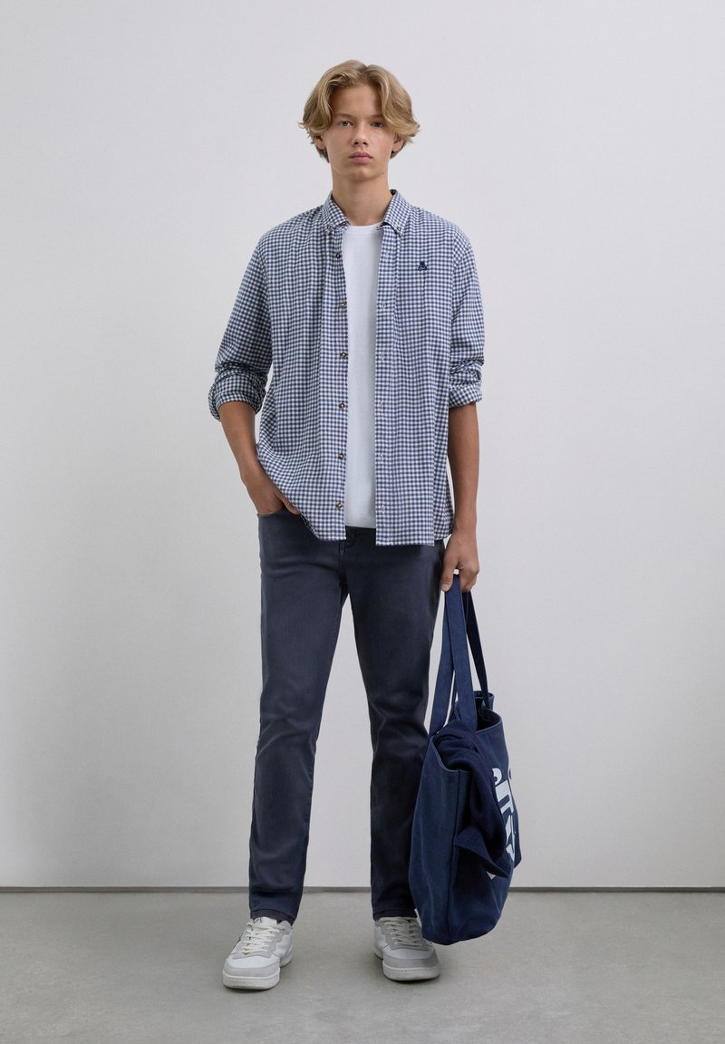 Joven vestido con una camisa a cuadros sobre una camiseta blanca, jeans oscuros, zapatillas blancas, sosteniendo una bolsa de tela azul marino con un suéter doblado dentro.