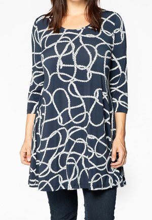 Laivastonsininen pitkähihainen tunika valkoisella köysikuvioinnilla, käytetty tummien housujen päällä, näkymä kaulasta reisiin.