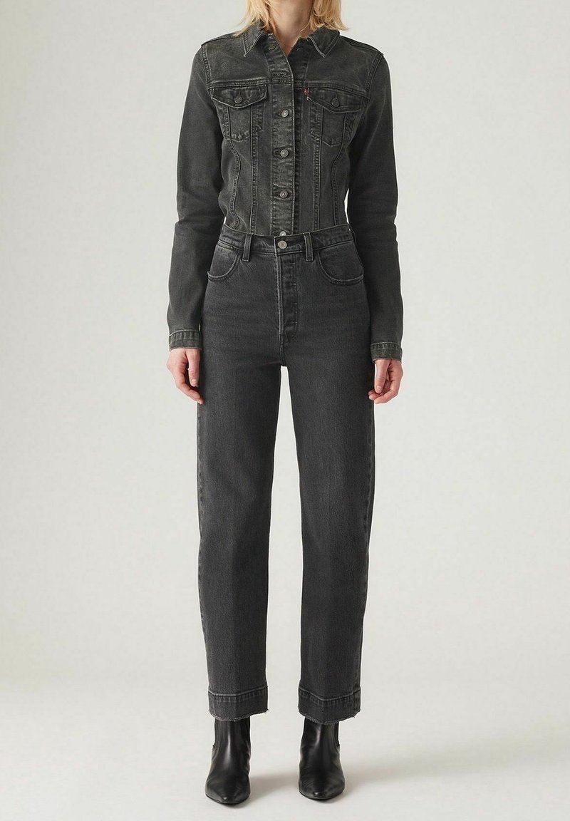 Femme portant une veste en jean noire ajustée et un jean noir taille haute, assortis à des bottines noires, debout sur un fond uni.
