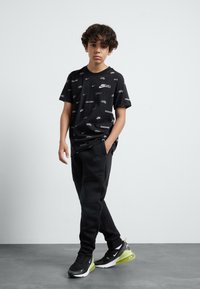 T-shirt graphique noir avec des logos Nike blancs, associé à un pantalon de survêtement noir et des baskets noires avec des accents verts et des semelles blanches.