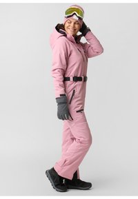 Pink ski suit with black accents, featuring a hood, zippered pockets, belt, and gray gloves. Worn with ski goggles and sturdy black boots.