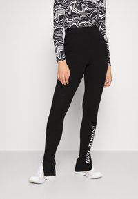 Black ribbed leggings with a high waistband. Features white text "KANI" on the left leg. Paired with white athletic shoes and a patterned top.