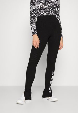 Black ribbed leggings with a high waistband. Features white text "KANI" on the left leg. Paired with white athletic shoes and a patterned top.