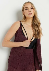 Burgunderröd stickad cardigan med ett spetsmönster, knappstängning och öppen front, lagrad över en svart topp med tunna remmar.