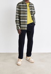 Grön- och vitrutig skjorta över en gul t-shirt, kombinerad med marinblå byxor och beige sneakers med vita accenter och subtila texturer.