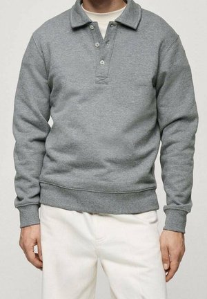 Mann trägt einen grauen Pullover mit langen Ärmeln und Kragen sowie eine weiße Hose, steht mit entspannten Armen an den Seiten.