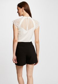 Haut blanc avec des manches courtes en dentelle et un dos en maille, présentant une fermeture en clé. Associé à un short noir, présenté sur un fond neutre.