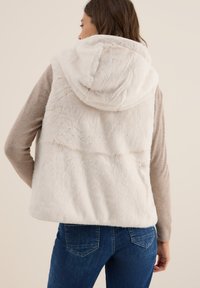 Vit fuskpälsväst med huva, med en mjuk textur och sömdetaljer, bärs över en beige topp och kombineras med blå jeans.