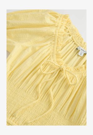 Keltainen pusero, jossa on pleated-tekstuuri, röyhelökaulus ja solmide tail. Taittuva vyötärö ja kevyt kangas. Merkkilogo näkyvissä.