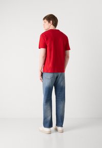 T-shirt en coton rouge à manches courtes, associée à un jean en denim bleu clair avec un délavage léger, et des baskets blanches. Design moderne et simple.