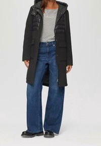 Abrigo largo negro con parte superior gris esponjosa, camisa a rayas debajo, pantalones vaqueros amplios azules y botas gruesas negras. Presenta cremallera y múltiples bolsillos.