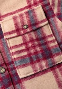 Chemise en laine beige avec un motif tartan rouge et bleu, comportant une poche à rabat et un grand bouton. Texture douce, col standard, couture visible.