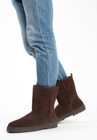 Botas de ante marrón hasta el tobillo con una textura suave, que presentan una punta redonda y una suela de goma resistente. Combinadas con unos jeans ajustados de mezclilla azul.