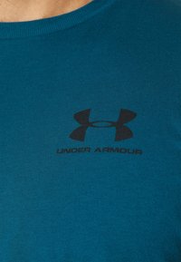 Tummansininen puuvillapaita pyöreällä kaula-aukolla, jossa on musta Under Armour -logo ja teksti ylärinnassa. Luja pinta.