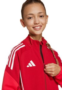 Giacca sportiva rossa con chiusura a zip, strisce bianche di accento sulle spalle e logo sul lato sinistro del petto. Tessuto sintetico liscio.