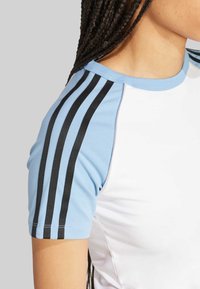 Košulja kratkih rukava s bijelim tijelom, svijetloplavim rukavima i crnim prugama. Ima okrugli izrez i glatku teksturu tkanine.