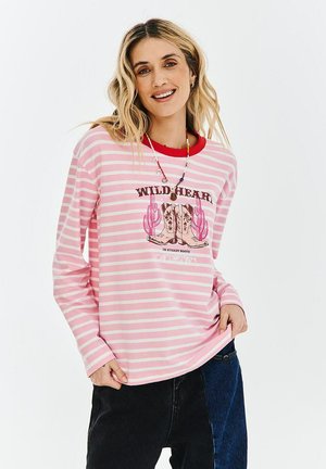 Uśmiechnięta kobieta z falującymi blond włosami nosi różowo-białą pasiastą bluzkę z długim rękawem z nadrukiem kowbojskich butów i kaktusa.