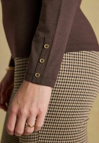 Brązowy długi rękaw z okrągłym dekoltem i detalami w postaci guzików w okolicy nadgarstka, w połączeniu z spodniami w wzór houndstooth.