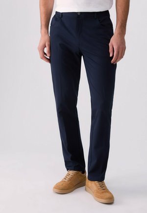 Homme portant un pantalon slim bleu marine foncé et des baskets en daim couleur sable, debout devant un fond uni.