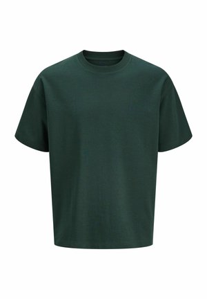Garçon debout, portant un t-shirt oversize vert foncé, un pantalon noir ample et des chaussures beiges sur un fond clair uni.