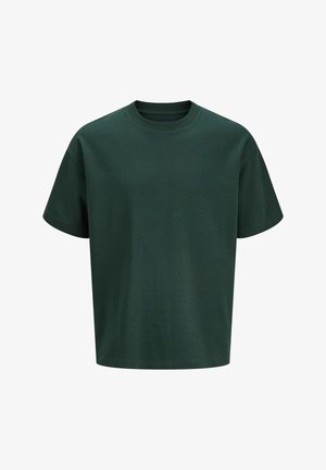Garçon debout, portant un t-shirt oversize vert foncé, un pantalon noir ample et des chaussures beiges sur un fond clair uni.