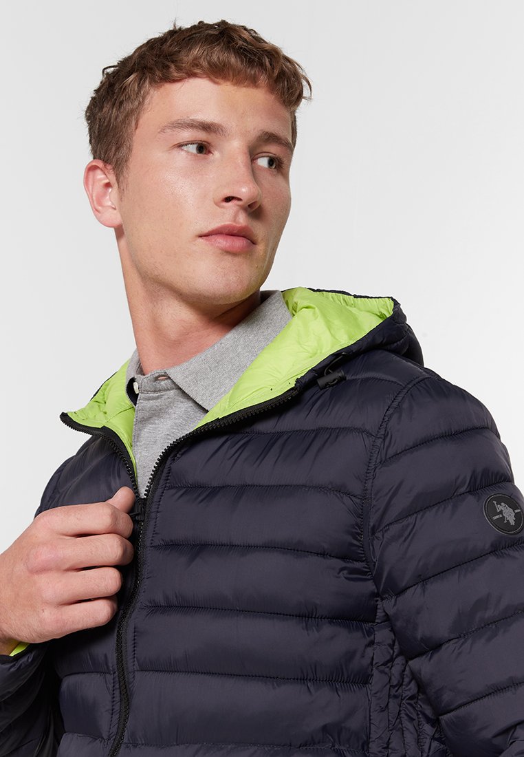 Marine pufferjack met een groene voeringskap, voorzien van horizontale stiksels, een ritssluiting en een logopatch op de mouw.