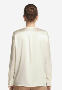 Långärmad skjorta i krämfärgad satin, med en bakrygg, subtila veckdetaljer och knäppta manschetter med sidofickor för ökad rörlighet.