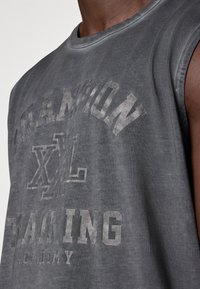 Tamsiai pilkas berankovė medvilninė marškinėlė su išblukusiu užrašu "CHAMPION XL TRAINING ACADEMY", su apvalia iškirpte ir laisvu siluetu.