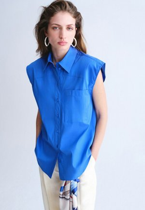 Mujer con camisa sin mangas de botones azul brillante, pantalones blancos, pendientes de aro plateados, de pie con las manos en los bolsillos contra un fondo claro.