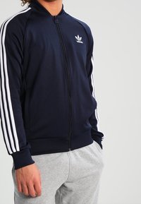 Giacca da track con zip di colore blu navy, con design a tre strisce bianche sulle maniche e logo. Tessuto morbido con polsini e vita a coste.