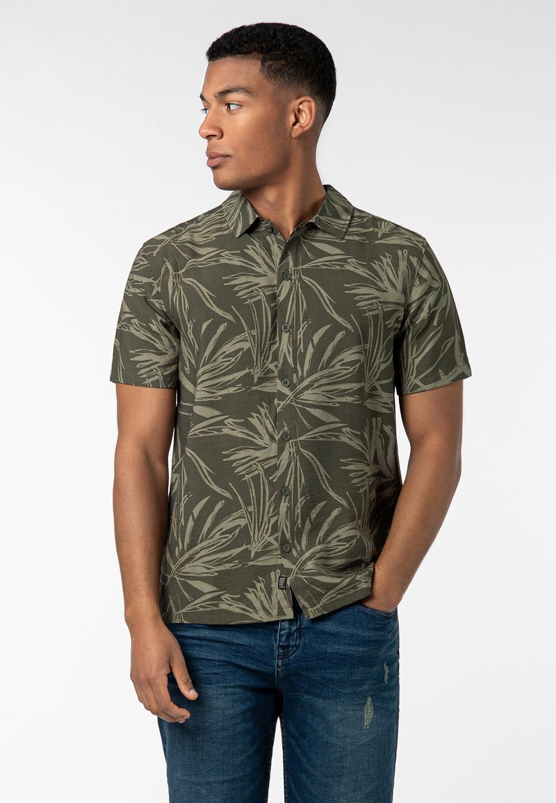 Kurzarmhemd aus olivgrünem Stoff mit einem blättrigen Muster. Verfügt über einen Kentkragen und einen geraden Saum, kombiniert mit blauen Jeans.