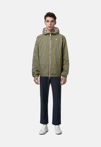 Giacca leggera verde oliva con cappuccio, zip frontale e dettagli a blocchi di colore; abbinata a pantaloni blu navy e sneakers bianche.