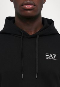 Felpa nera con cappuccio realizzata in tessuto morbido, con cappuccio regolabile e logo bianco EA7 Emporio Armani sul petto.