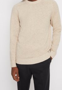 Beige stickad tröja med ribbad rundhals och ärmslut. Tyget har en mjuk textur och ett subtilt melerat mönster. Kombinerat med mörka byxor.