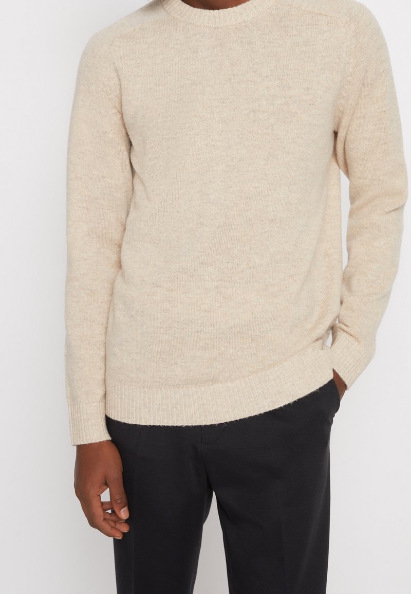 Beige stickad tröja med ribbad rundhals och ärmslut. Tyget har en mjuk textur och ett subtilt melerat mönster. Kombinerat med mörka byxor.