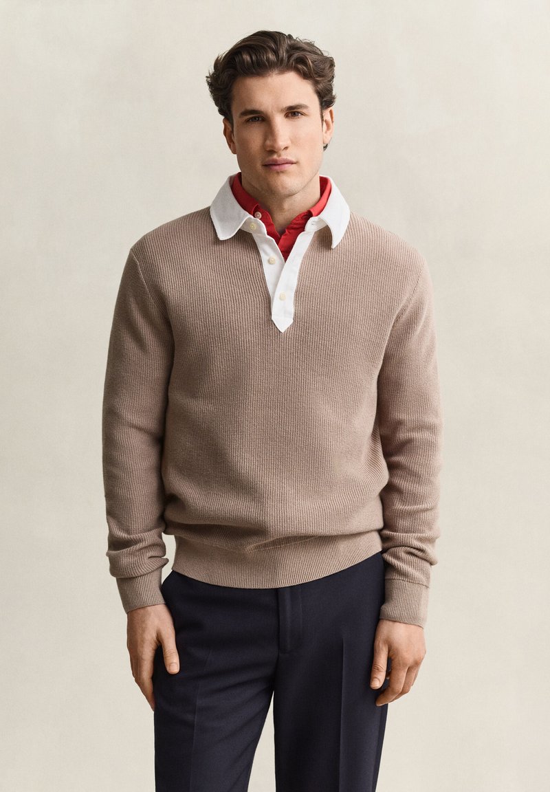 Man wearing beige knit sweater over white collared shirt and red undershirt, standing against plain light background.